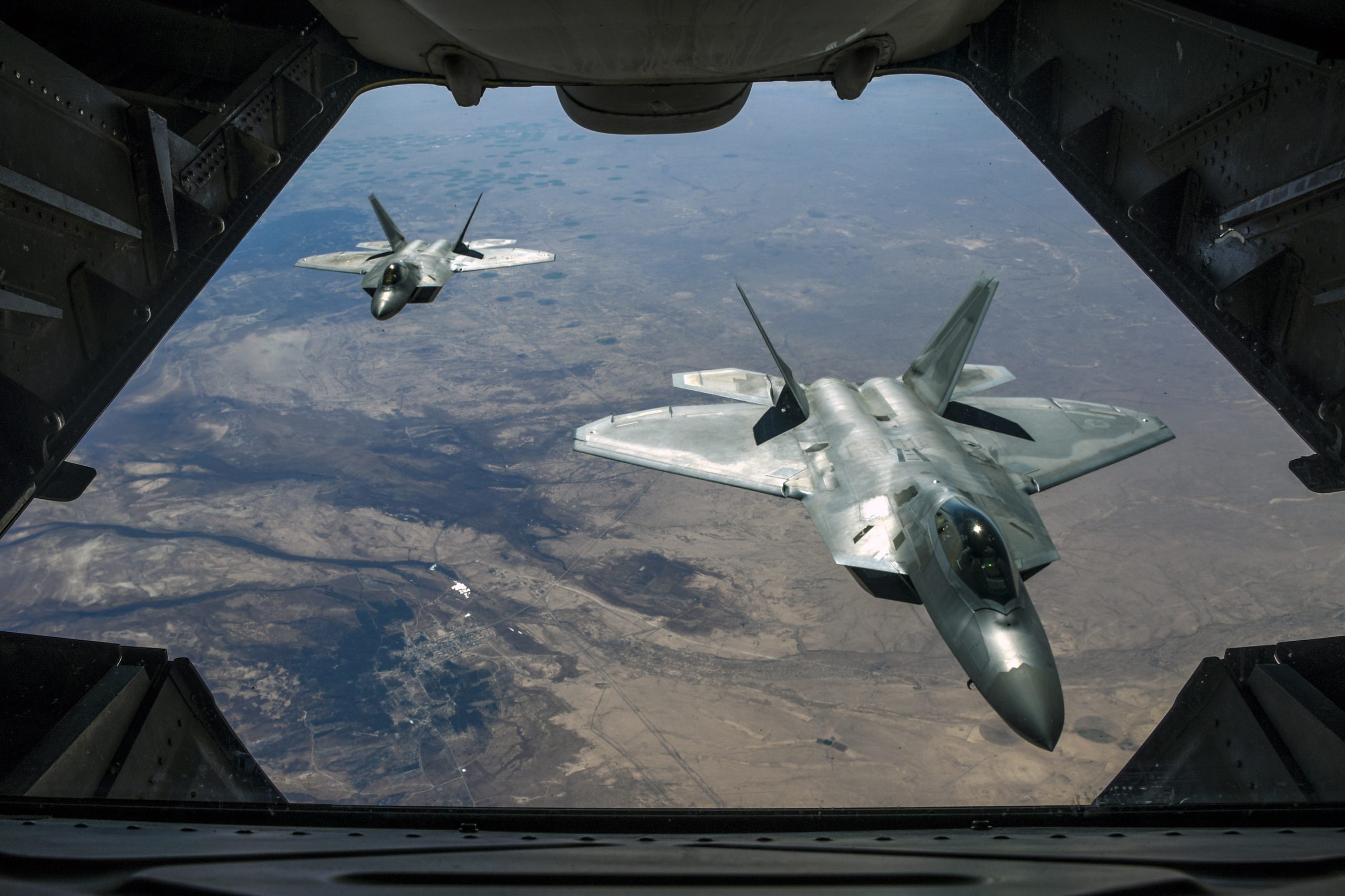 Two fighter jets, viewed from an opening in a third aircraft in front of them, fly over brownish terrain.