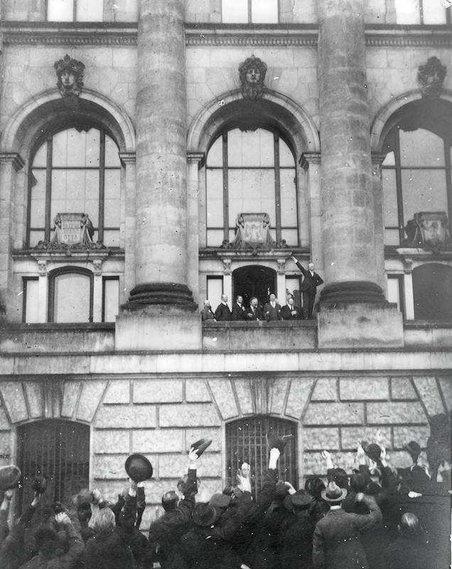 - Ausrufung Republik Scheidemann 1 Philipp Scheidemann, bras levé, proclame la République allemande depuis un balcon du Reichstag, le 9 novembre 1918 — Guillaume II vient de renoncer au trône le jour même, mettant fin à près de cinquante ans de monarchie. À quelques centaines de mètres de là, Karl Liebknecht proclame, ce même jour, une République socialiste rivale : une nouvelle époque commence, mais deux Allemagnes s’en disputent aussitôt le sens — Photo Erich Greiser / Wikipedia