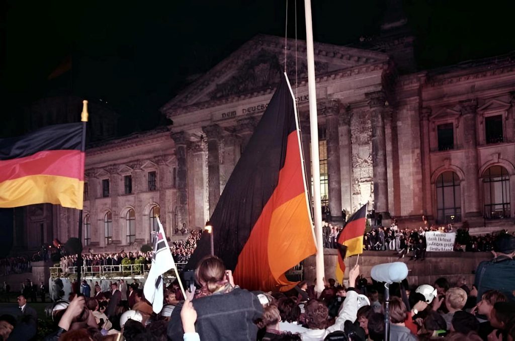 Trois mois plus tard, dans la nuit du 2 au 3 octobre 1990, un immense drapeau noir-rouge-or se hisse devant le Reichstag, sous les projecteurs, face à une foule immense. L’Allemagne n’est plus un pays occupé, ni un pays divisé, ni un pays sous tutelle. Elle est, pour la première fois depuis 1945, un État comme les autres — Photo Bundesarchiv / Grimm